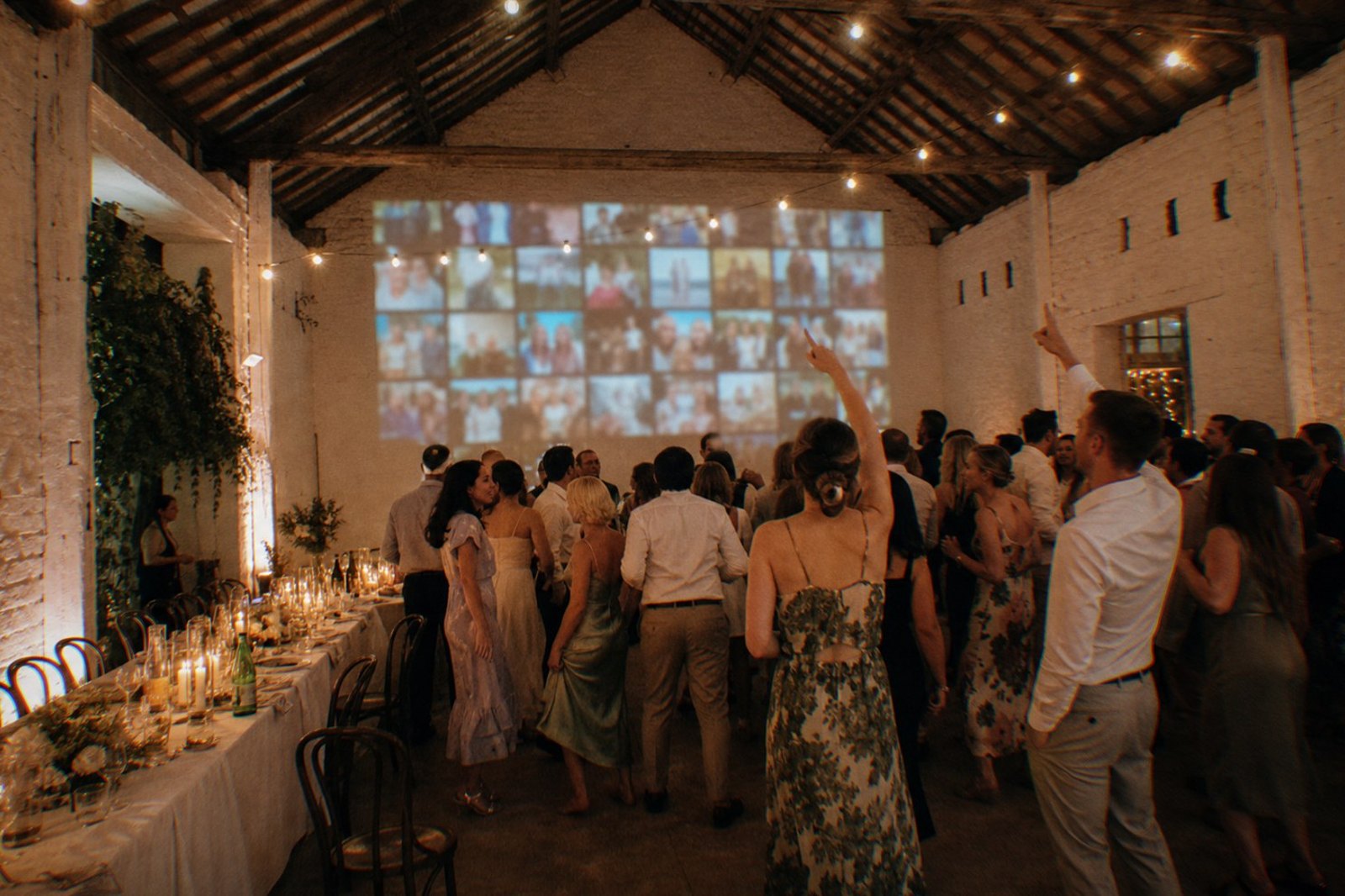 Una recepción de boda en un granero de paredes encaladas de noche, con un muro de fotos en vivo proyectado en la pared del fondo, invitados bailando y señalándolo