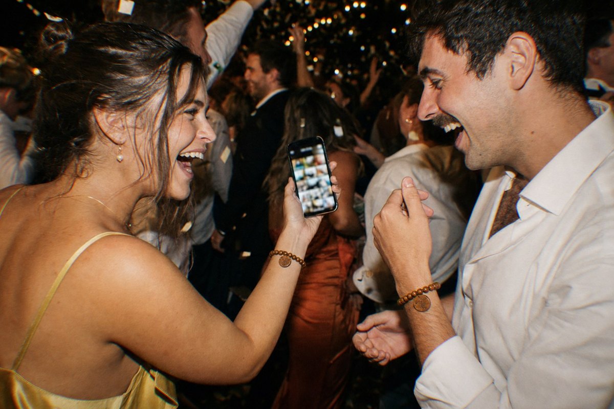 Two laughing guests on a dark dance floor looking at a glowing photo gallery on a phone, both wearing wooden bead bracelets with engraved tags