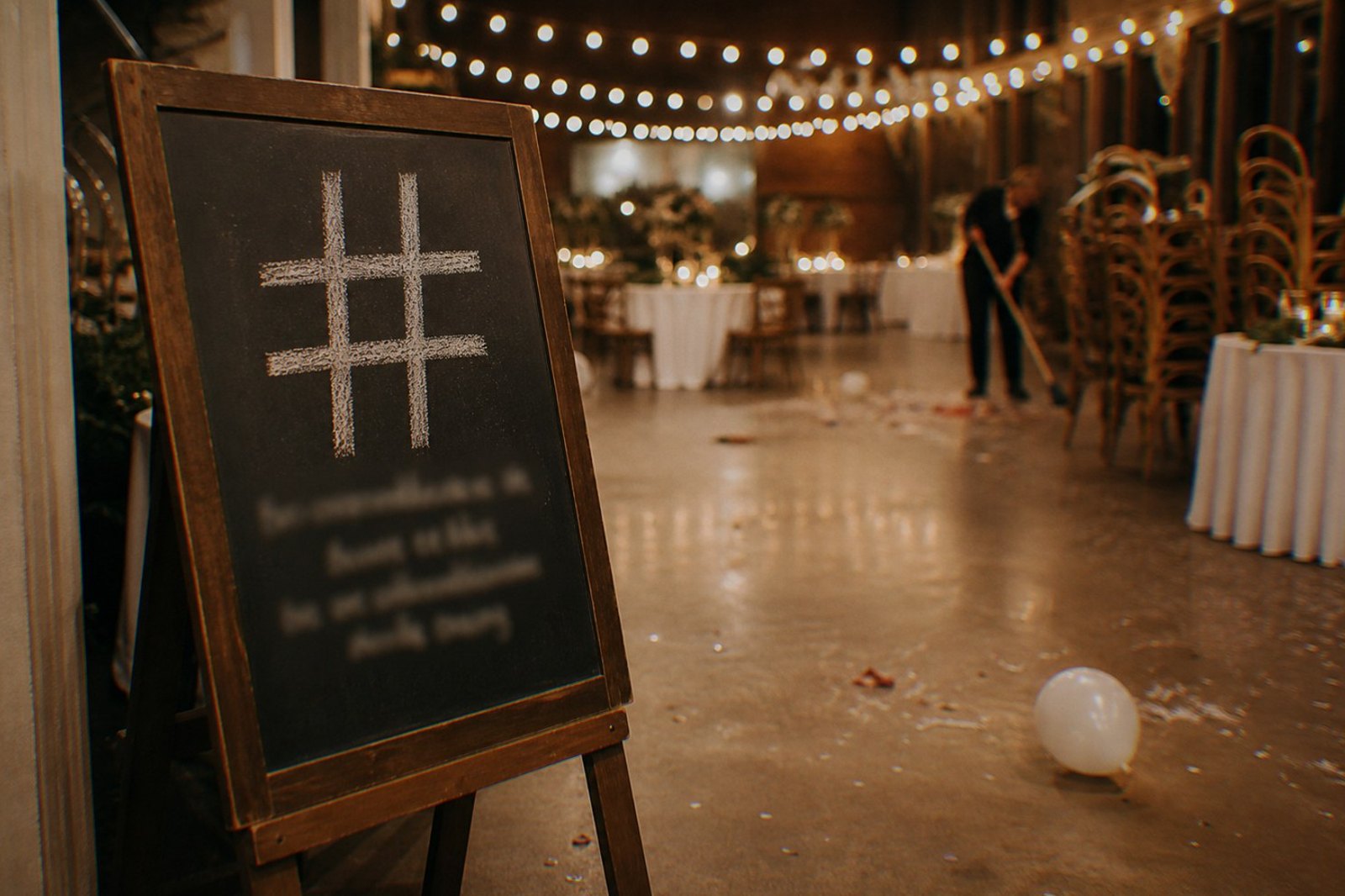 A chalkboard easel sign with a large chalk hashtag symbol leaning askew at an emptying wedding reception at the end of the night, stacked chairs and string lights behind